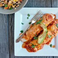 Two enchiladas with red sauce topped with avocado on a white plate with a fork beside them.