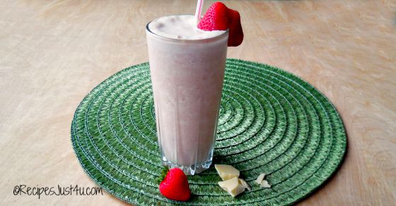 A tropical white chocolate smoothie garnished with a strawberry, in a glass on a round green placemat, with a strawberry and white chocolate pieces on the placemat beside it.