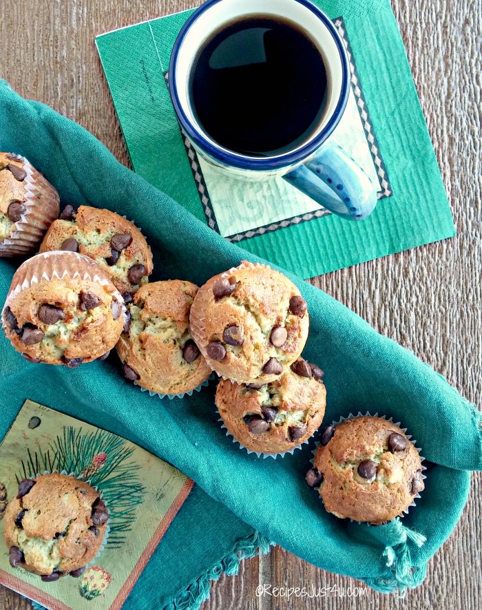 Seven banana chocolate chip muffins in a long basket lined with a green cloth beside a cup of black coffee.
