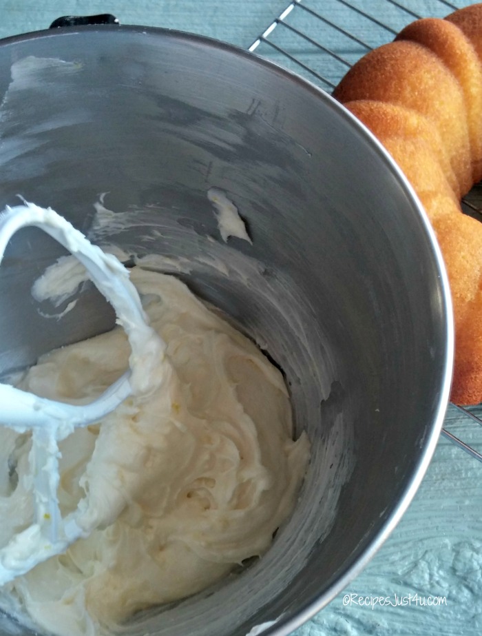 Lemon buttercream frosting in a mixing bowl with beater next to a cake.
