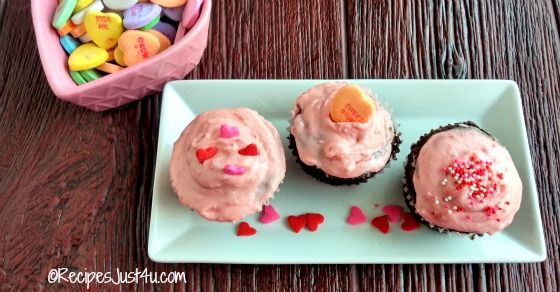 Three strawberry Valentine cupcakes on a plate next to a small heart shaped bowl of conversation hearts.