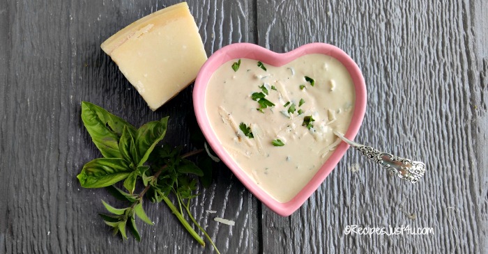 Parmesan herb dressing in a pink heart shaped bowl with spoon, fresh herbs and block of cheese.