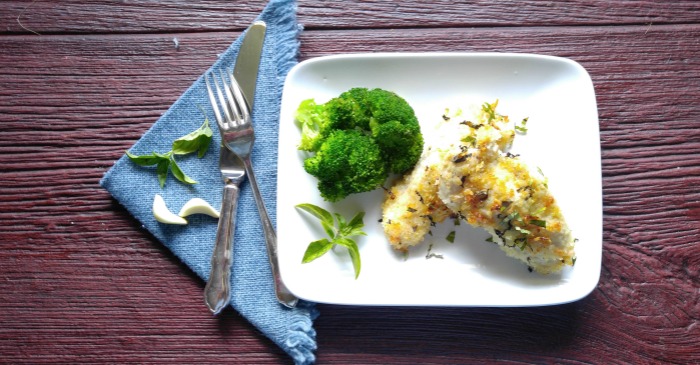 Oven baked Parmesan chicken tenders and broccoli on a white plate with a blue napkin and cutlery on it.