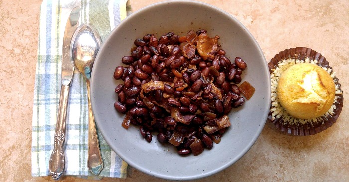 A bowl of crock pot Boston baked beans with a cornbread muffin to the right of the bowl, and cutlery on a napkin to the left of it.