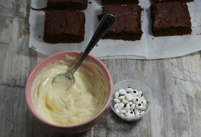 Melted white chocolate in a bowl with edible eyes near some Halloween brownies.