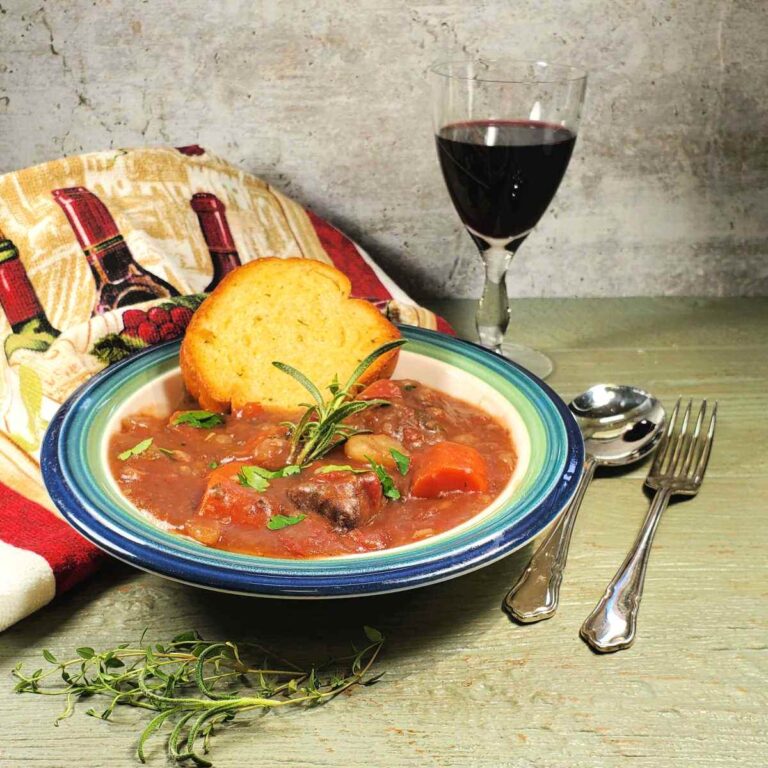 A bowl of beef stew and garlic bread sitting next to a wine themed towel and cutlery and a glass of red wine.