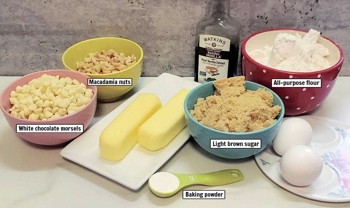 Macadamia nuts, white chocolate morsels, vanilla extract, butter, flour, brown sugar, eggs, and baking powder on a counter top.