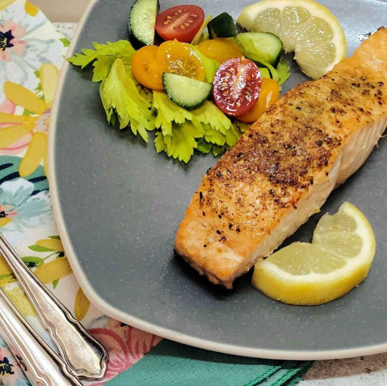 Baked salmon recipe, lemon pepper seasoning on top with salad and lemon slices on a gray plate.