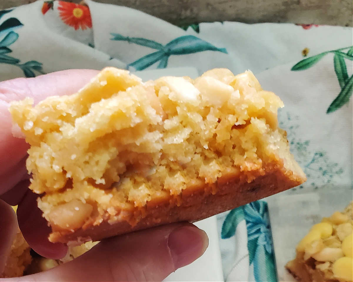A woman's hand holding a white chocolate macadamia slice over a flowered napkin with a bite out of the bar.