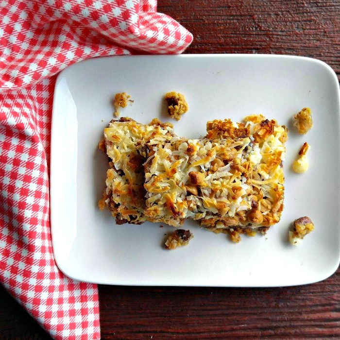 Two slices of 7 layer bars on a white plate sitting on a red and white checked napkin.