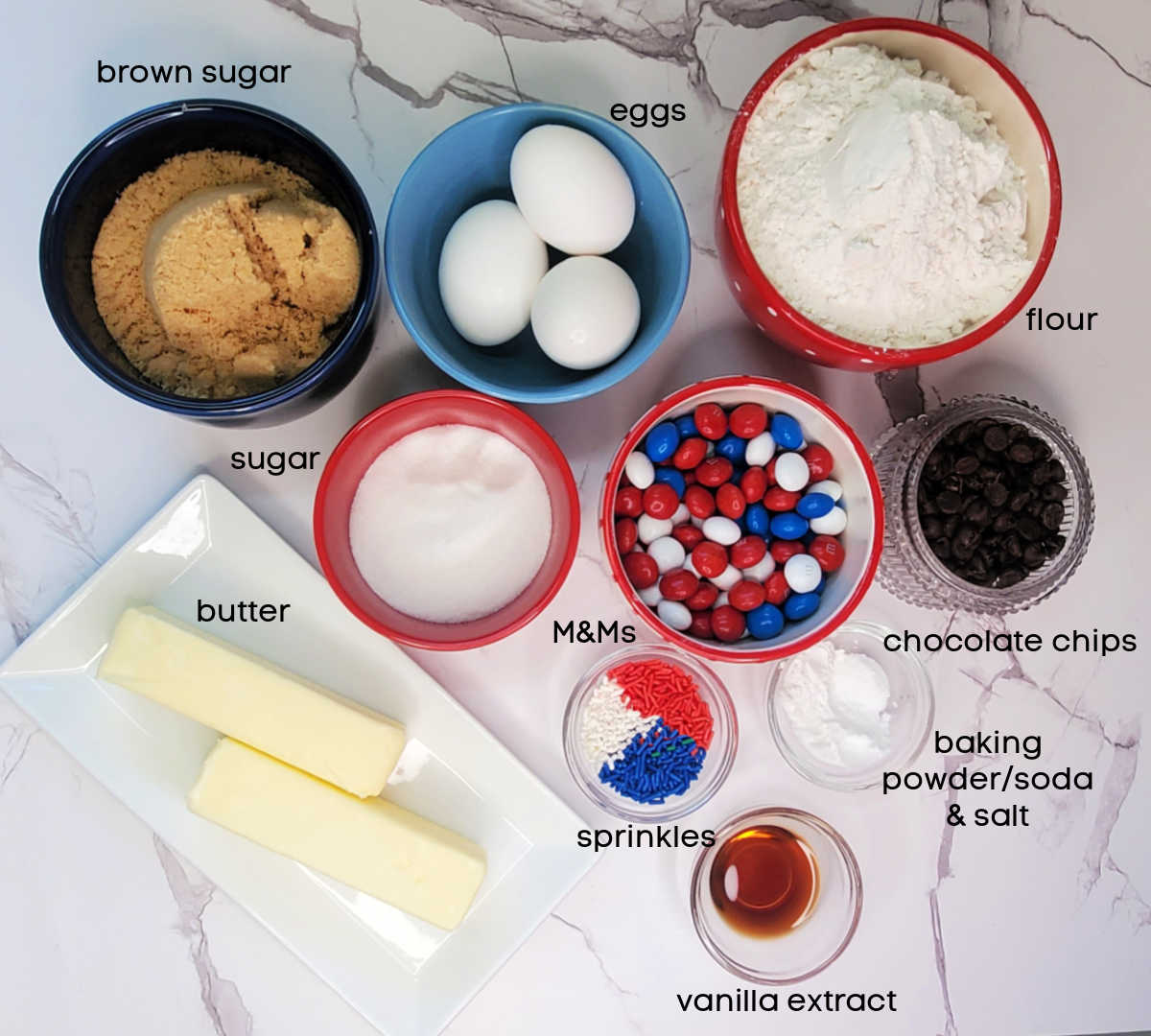 Bowls on a marble countertop with sugar, eggs, flour, M&Ms, chocolate chips, butter, sprinkles, vanilla, and baking soda, baking powder and salt.
