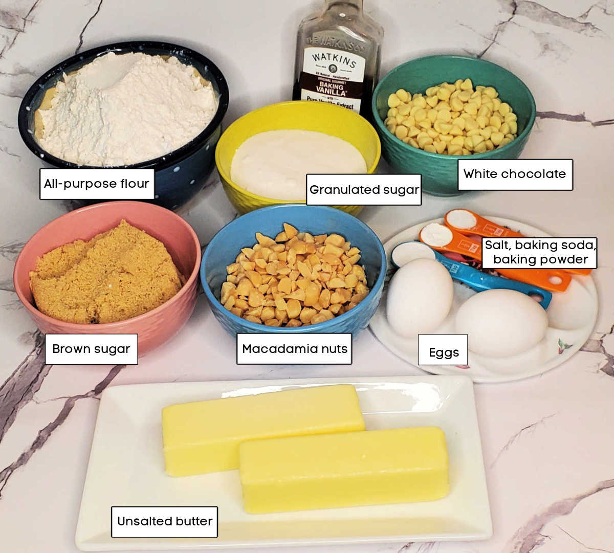 Butter, brown sugar, macadamia nuts, eggs, baking soda, baking powder, salt, vanilla, flour, sugar and white chocolate in bowls on a marble countertop. Small text boxes label all the ingredients.