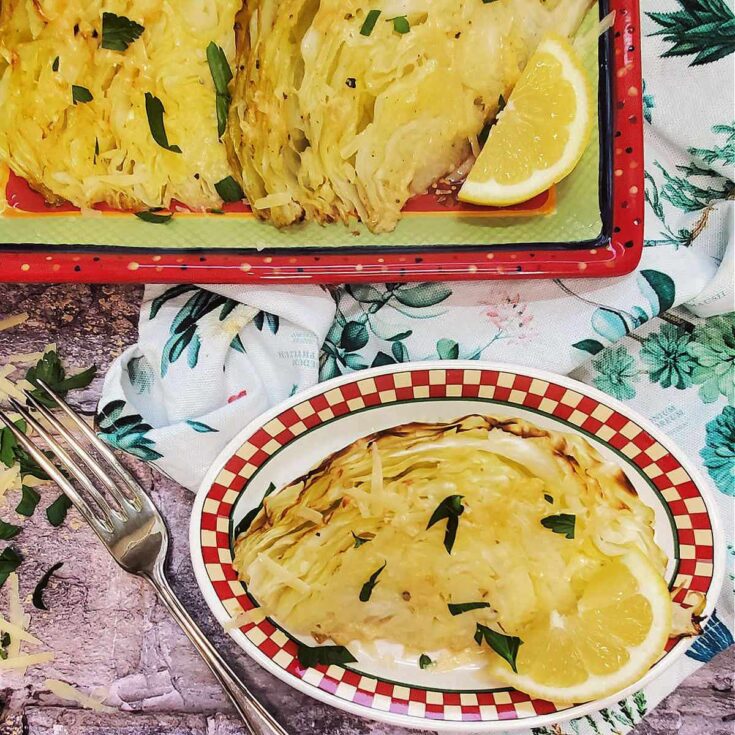 A red and white plate with a wedge of roasted cabbage with lemon sitting on a flowered towel next to a serving dish with roasted cabbage wedges on it.