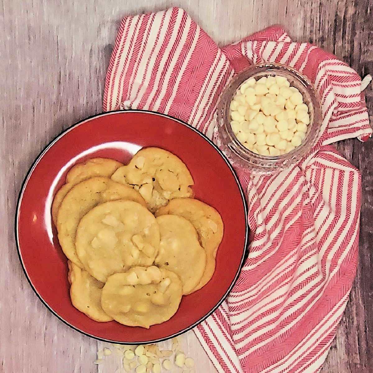 A red plate with macadamia nut white chocolate cookies sitting near a bowl of white chocolate chips and a red and white tea towel.