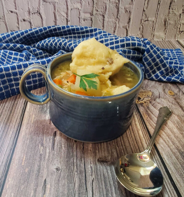 Blue bowl with a bowl of chicken dumplings with Bisquick next to a blue towel and soup spoon.