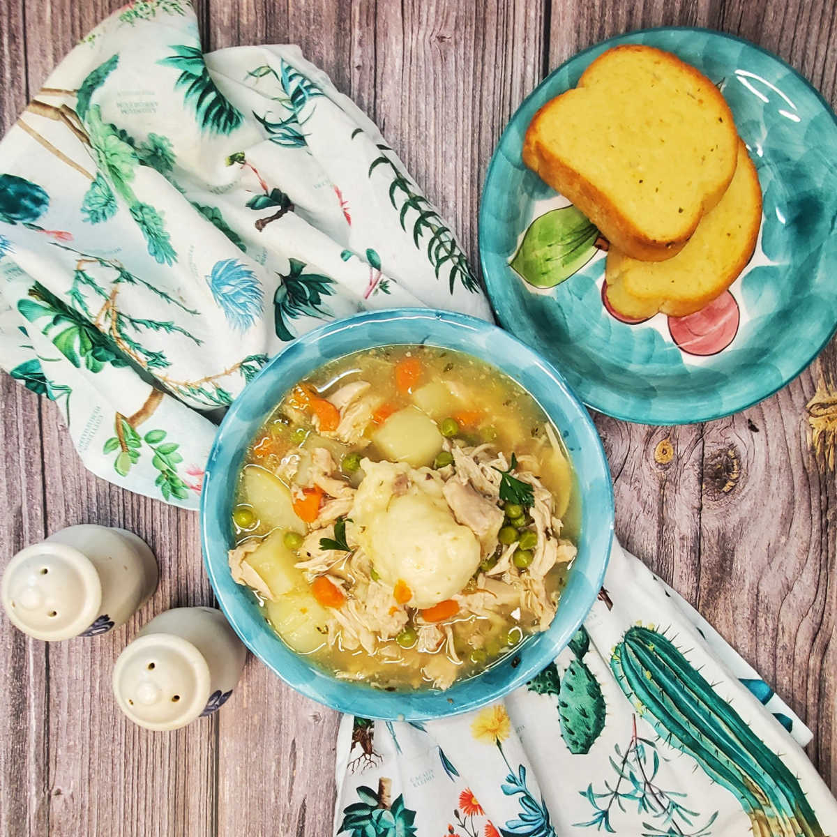 Serving chicken and dumplings A bowl of chicken and dumplings on a flowered towel sitting next to salt and pepper sharkers and two slices of garlic bread.