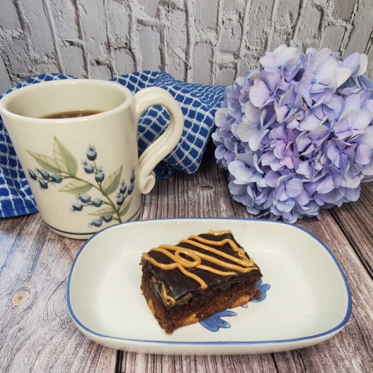 A chocolate peanut butter brownies recipe on a white plate in front of a blue hydrangea flower, blue cloth and cup of coffee.