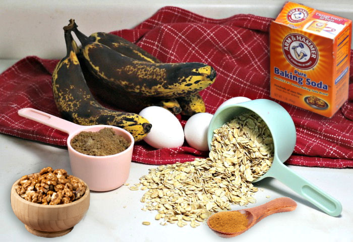 Bananas, eggs, baking soda, brown sugar, rolled oats, walnuts and cinnamon on a counter with a red towel.