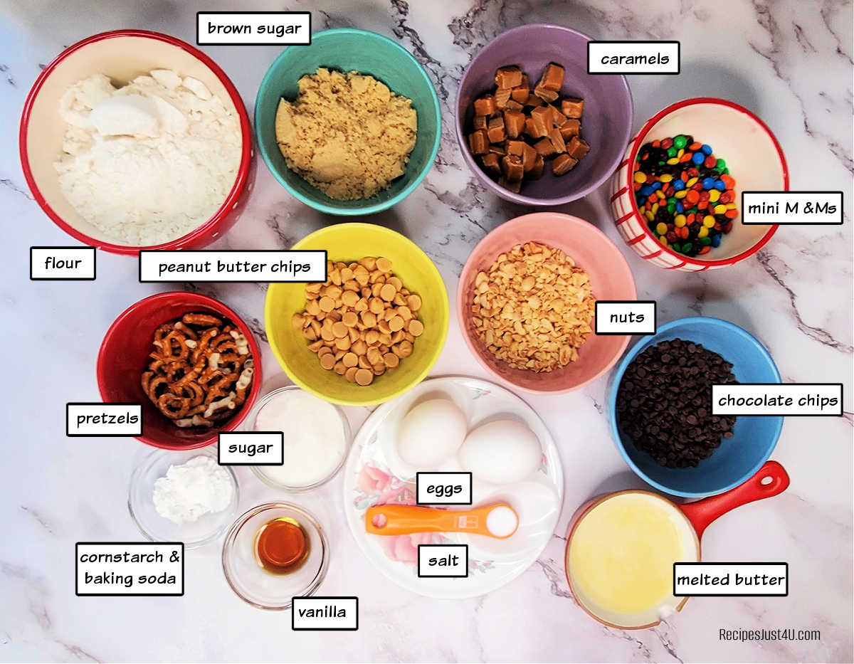 ingredients for making kitchen sink bars in bowls on a marble counter. Bowls contain flour, sugar, salt, eggs, butter, baking soda, cornstarch, vanilla and lots of fun mix ins.