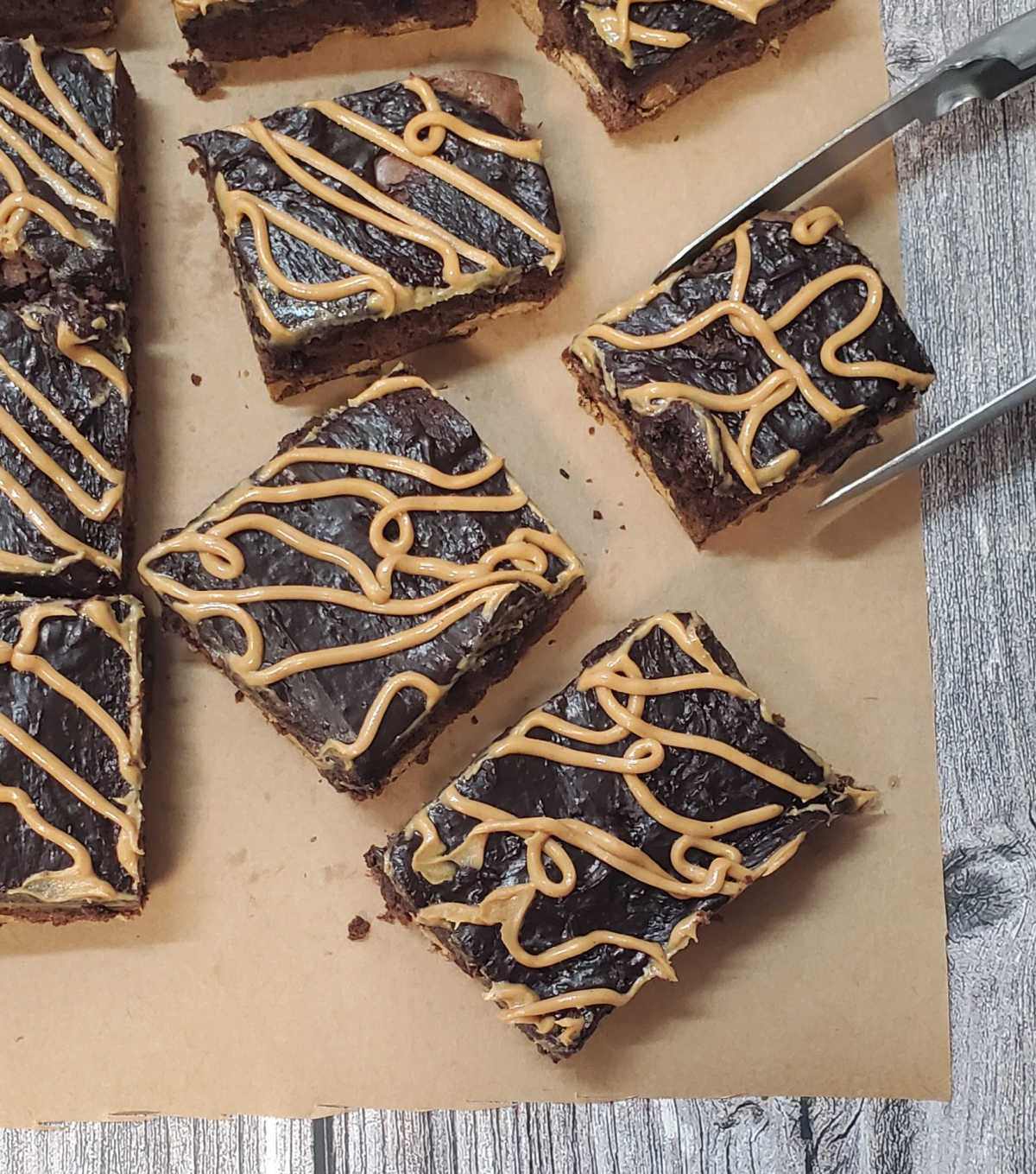 Chocolate peanut butter brownies on brown paper cut into squares with a pair of tongs removing one brownie.