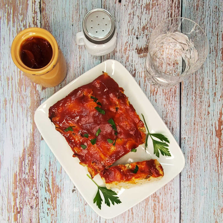 A turkey veggie meatloaf on a white plate with parsley and tomatoes, surrounded by salt, water and BBQ sauce.