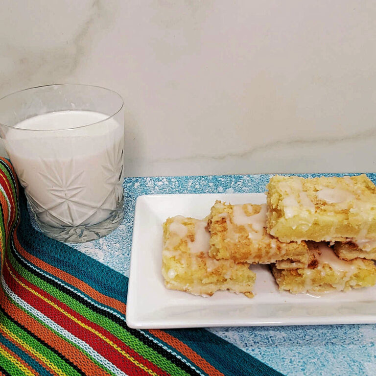 Lemon bars dessert recipe on a white plate next to a glass of milk and colorful striped towel.