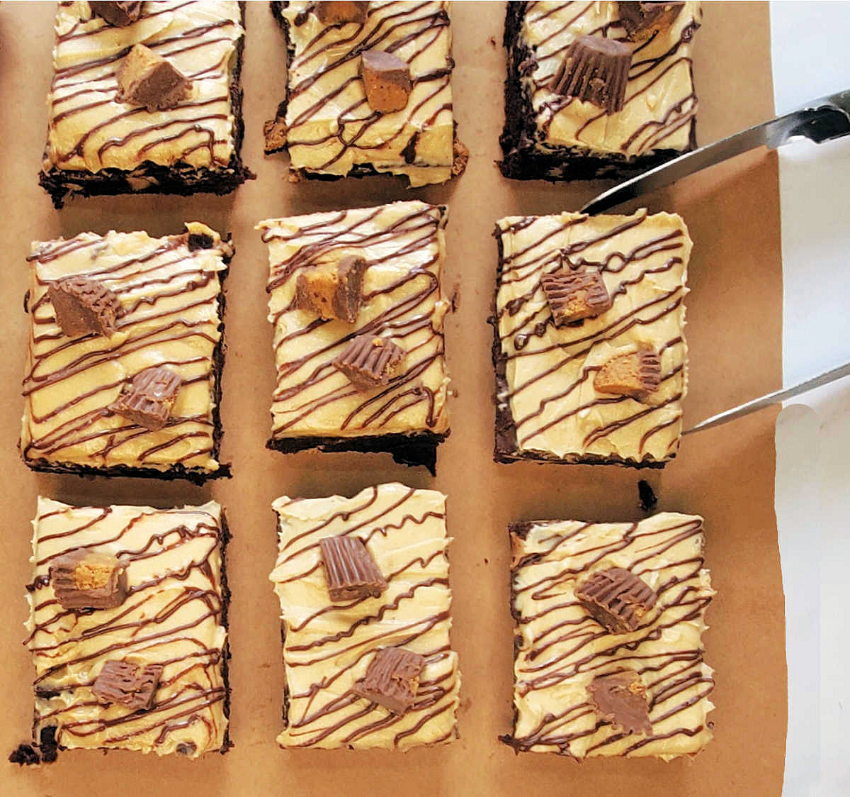 Brownies with peanut butter frosting on brown paper next to a pair of tongs trying to pick up a brownie.