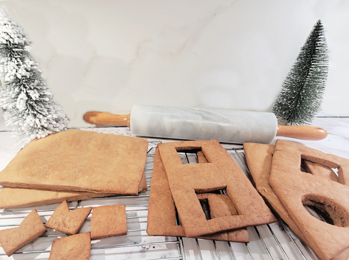 Cool the gingerbread pieces before building the house. Gingerbread house pieces cooling on wire racks on a countertop in front of a rolling pin and 2 small faux Christmas trees.