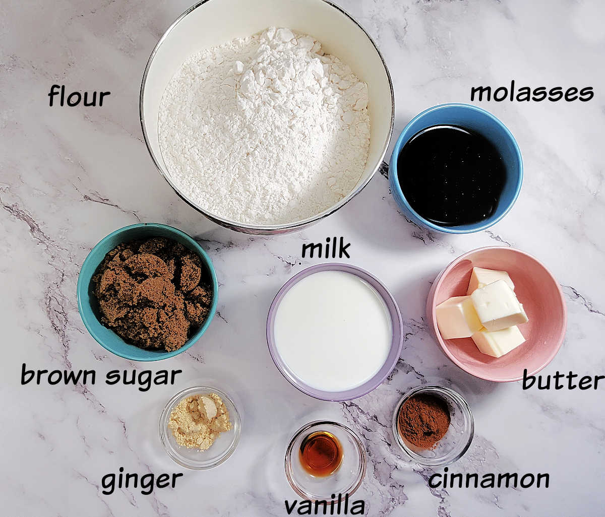 Gather your ingredients before you start. Bowls with labeled ingredients for gingerbread house dough on a marble counter, including flour, molasses, brown sugar, milk, butter, ginger, vanilla and cinnamon.