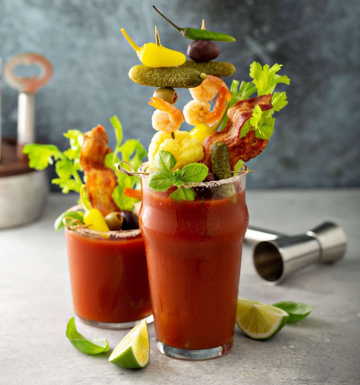 Two bloody Marys with elaborate cocktail garnishes on them, with limes beside the glasses and bar tools in the background.